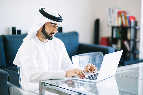 A Muslim man in traditional white attire working on a laptop at home